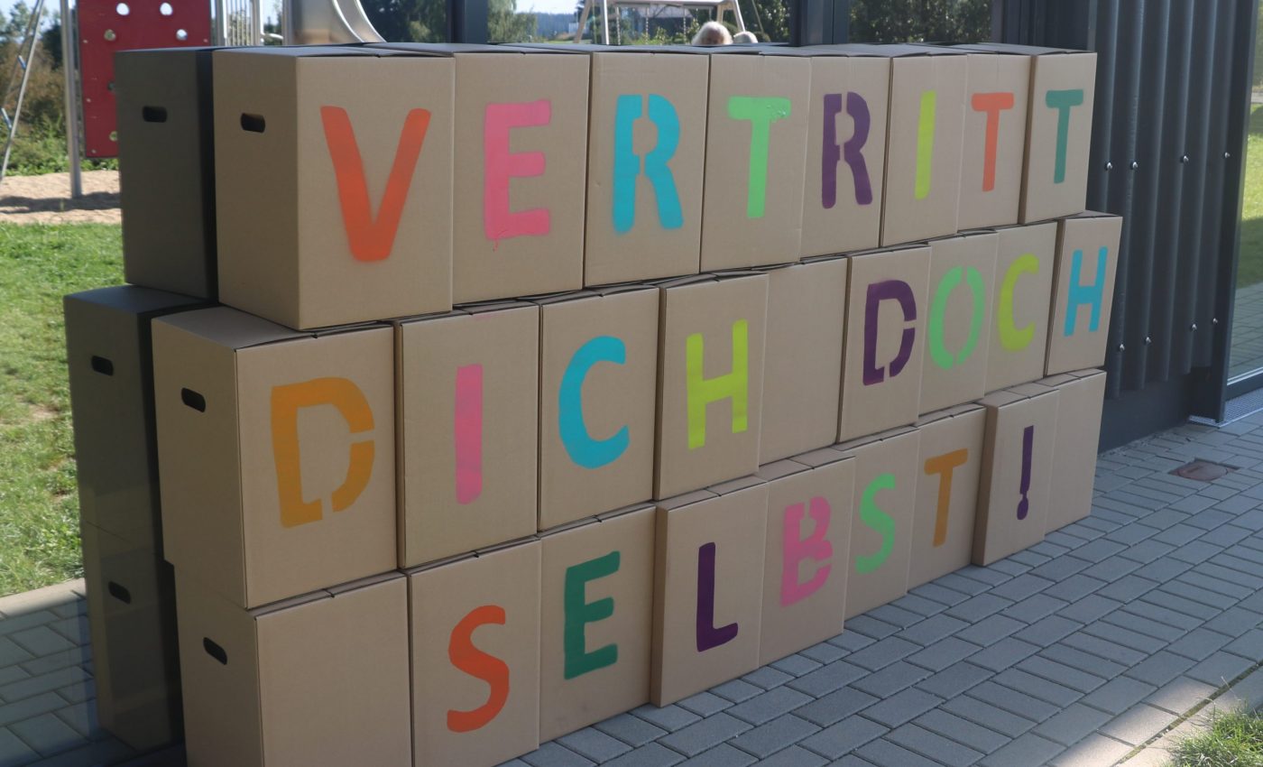 Auf diesem Bild sind Kartons zu sehen. Auf jedem Karton steht ein Buchstabe. Zusammen ergeben sie den Aufruf: Vertritt dich doch selbst!